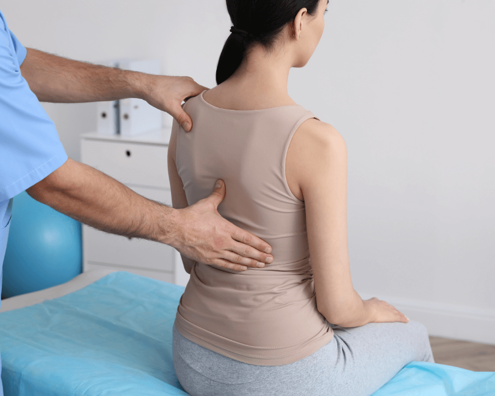 A healthcare professional assesses a patient's back during a physical therapy session, with treatment equipment and furnishings in the background.