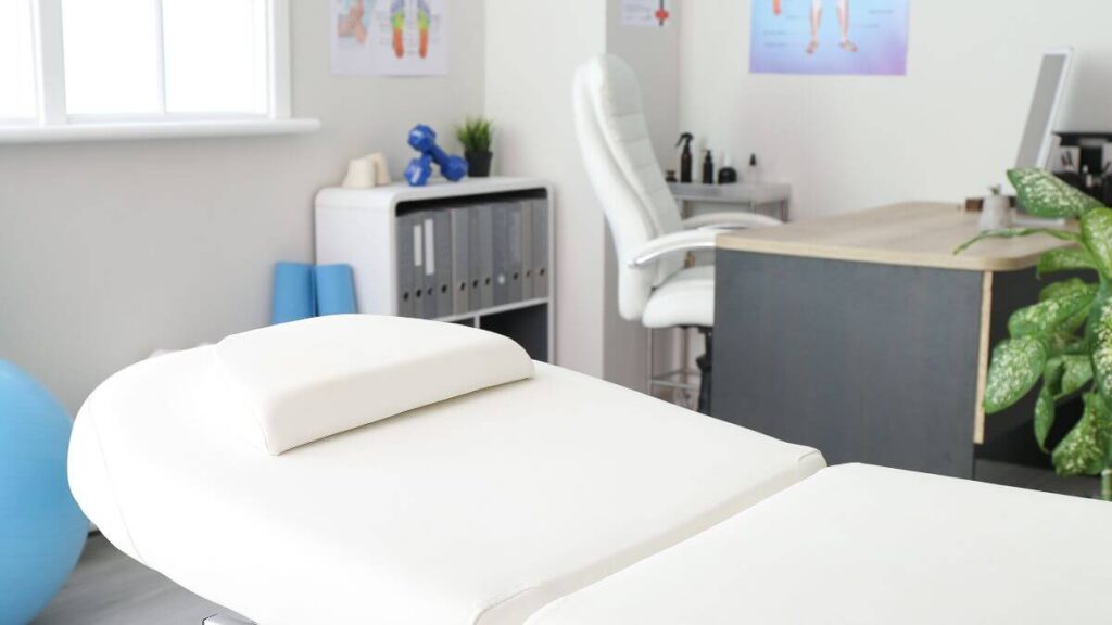 A clean treatment room with a white massage table, a desk, a chair, and exercise equipment, including a blue exercise ball and dumbbells.