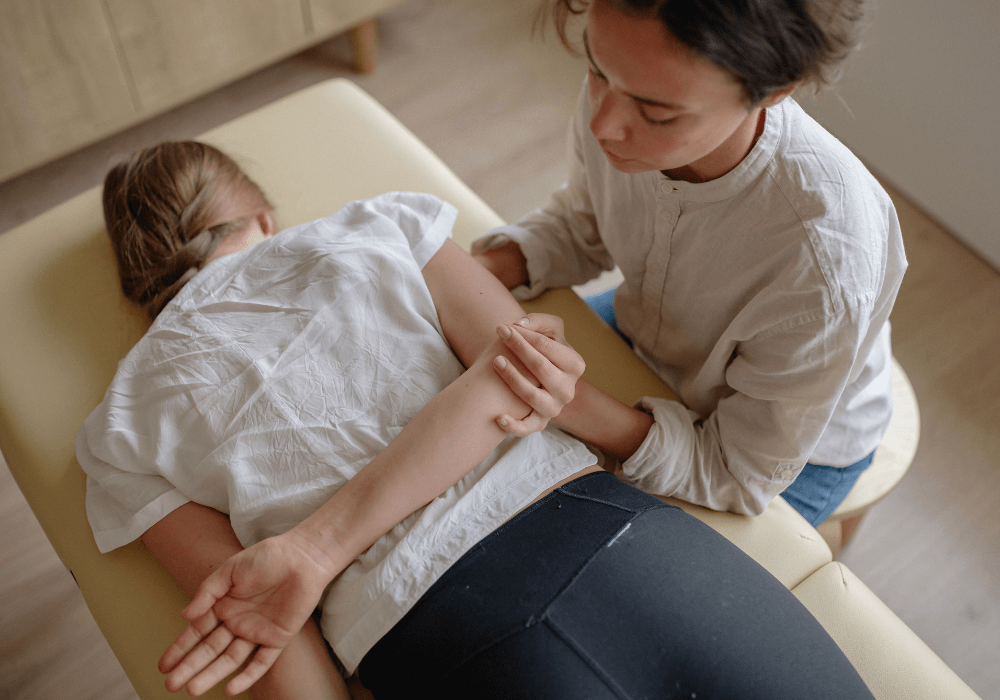 A person receiving a therapeutic treatment lies on a massage table, with a practitioner gently manipulating their arm.