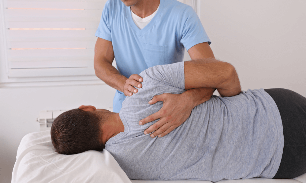 A therapist gently manipulates the shoulder of a client lying face down on a treatment table in a bright therapy room.