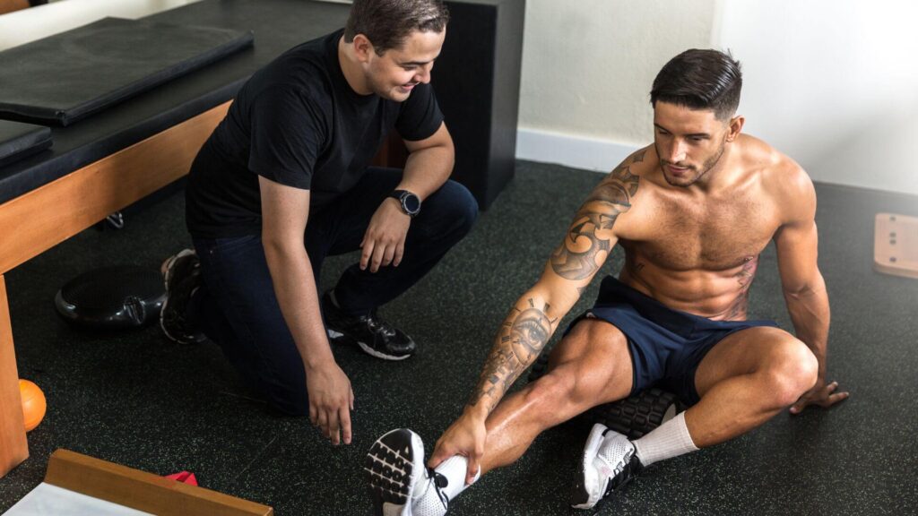 A shirtless man with tattoos stretches while sitting on a gym mat, receiving guidance from another person who is kneeling nearby.